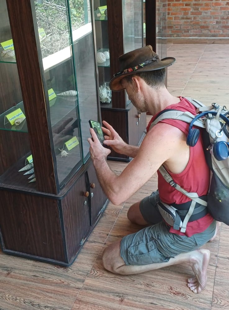 Young woman in a pink shirt explains something to two adults near a tented area with stairs; one listener wears a colorful cover-up, the other a teal tank top.