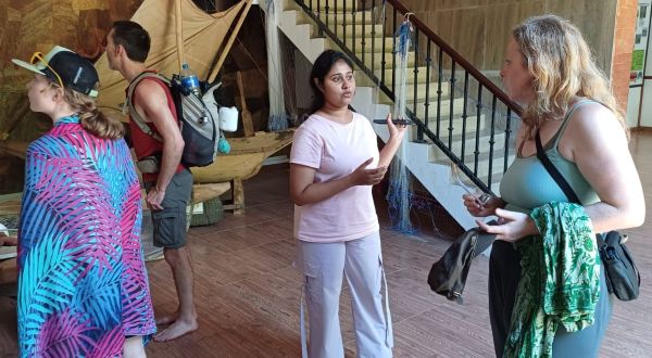 Young woman in a pink shirt explains something to two adults near a tented area with stairs; one listener wears a colorful cover-up, the other a teal tank top.