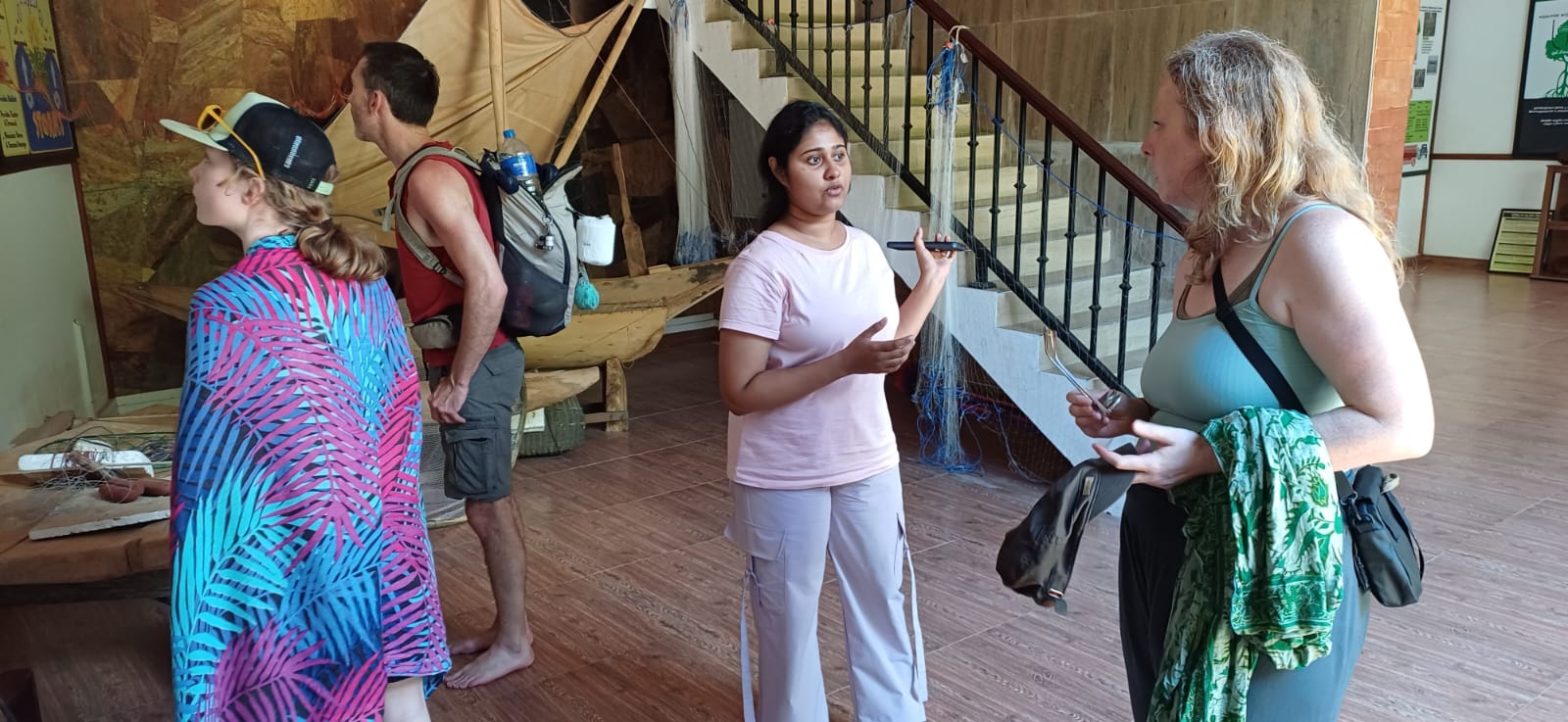 People chat indoors near a wooden boat exhibit with nets and gear in the background.