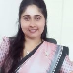 Smiling woman wearing a pink saree with a black border, seated in an office chair indoors.