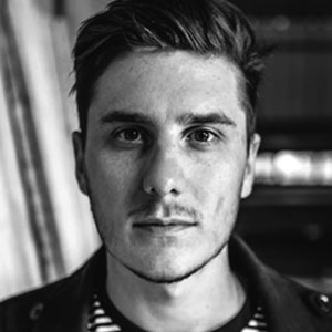 Close-up portrait of a young man with dark hair and a neutral expression, looking directly at the camera in black and white.
