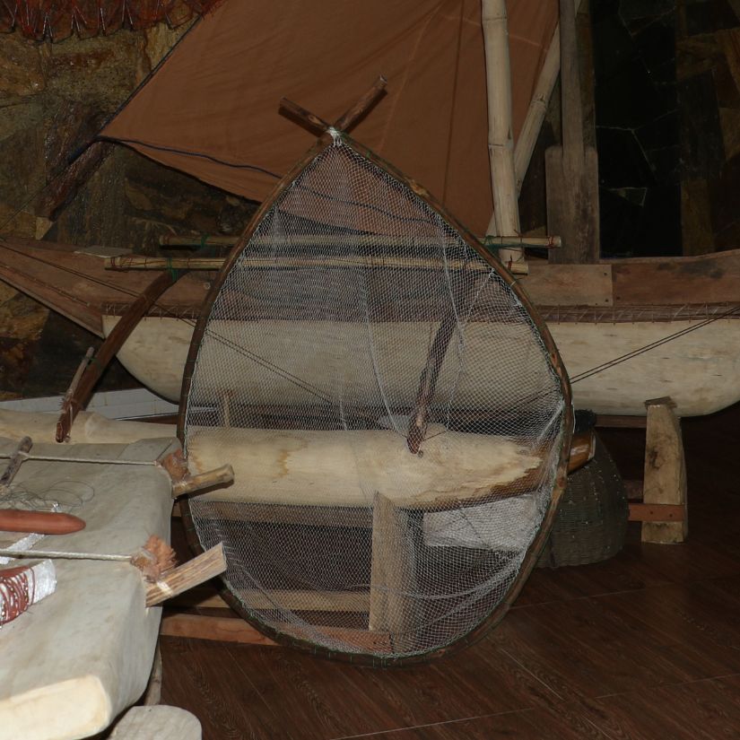 Round wicker fish trap with mesh netting and wooden supports resting on a wooden floor beside a boat in a cluttered workshop.