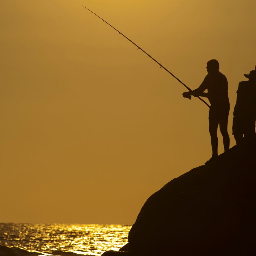 Two silhouetted fishermen stand on a rocky ledge, fishing into the golden sunset over the water.