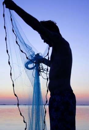 Silhouette of a person on a beach at sunset, holding up a translucent fishing net over their head over calm water.