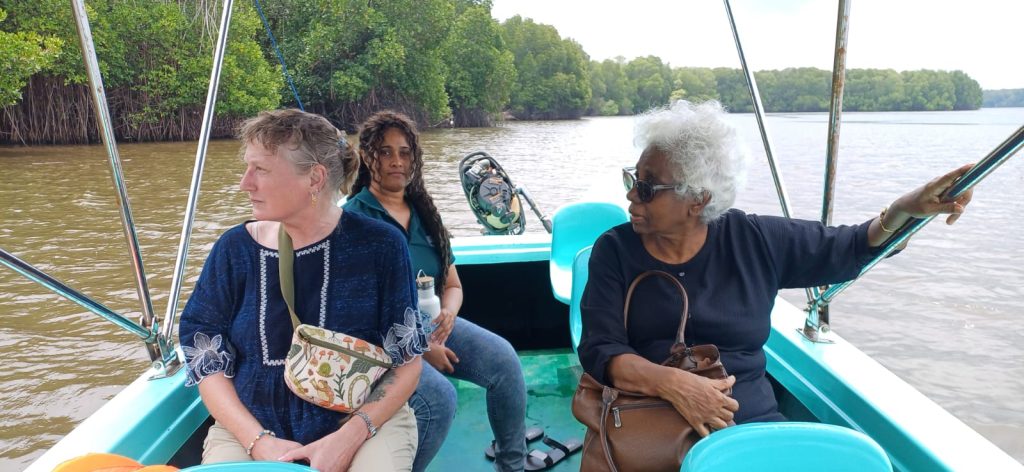 Three women ride in a small turquoise boat on a brown river, with mangroves in the background. The woman on the left wears a blue embroidered top and a patterned crossbody bag.