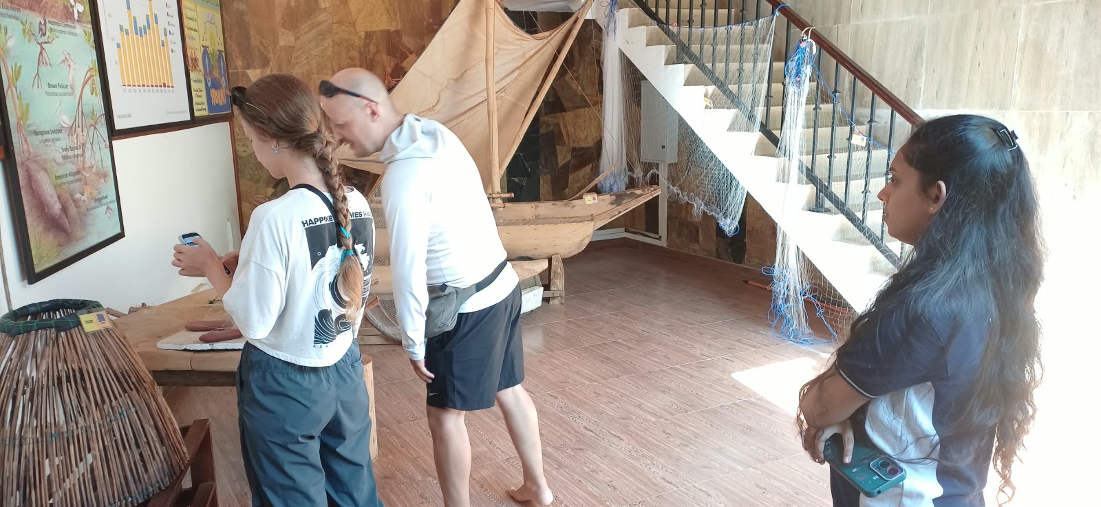 Three visitors in a museum examine a wooden boat model beside nautical posters on the wall.
